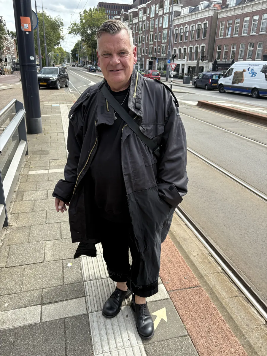 Man in a long black coat and leather shoes at a tram stop.