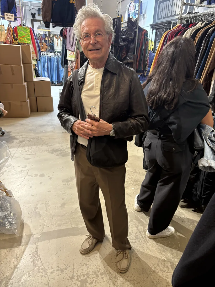 Older man in a leather jacket and brown trousers at a market stall.